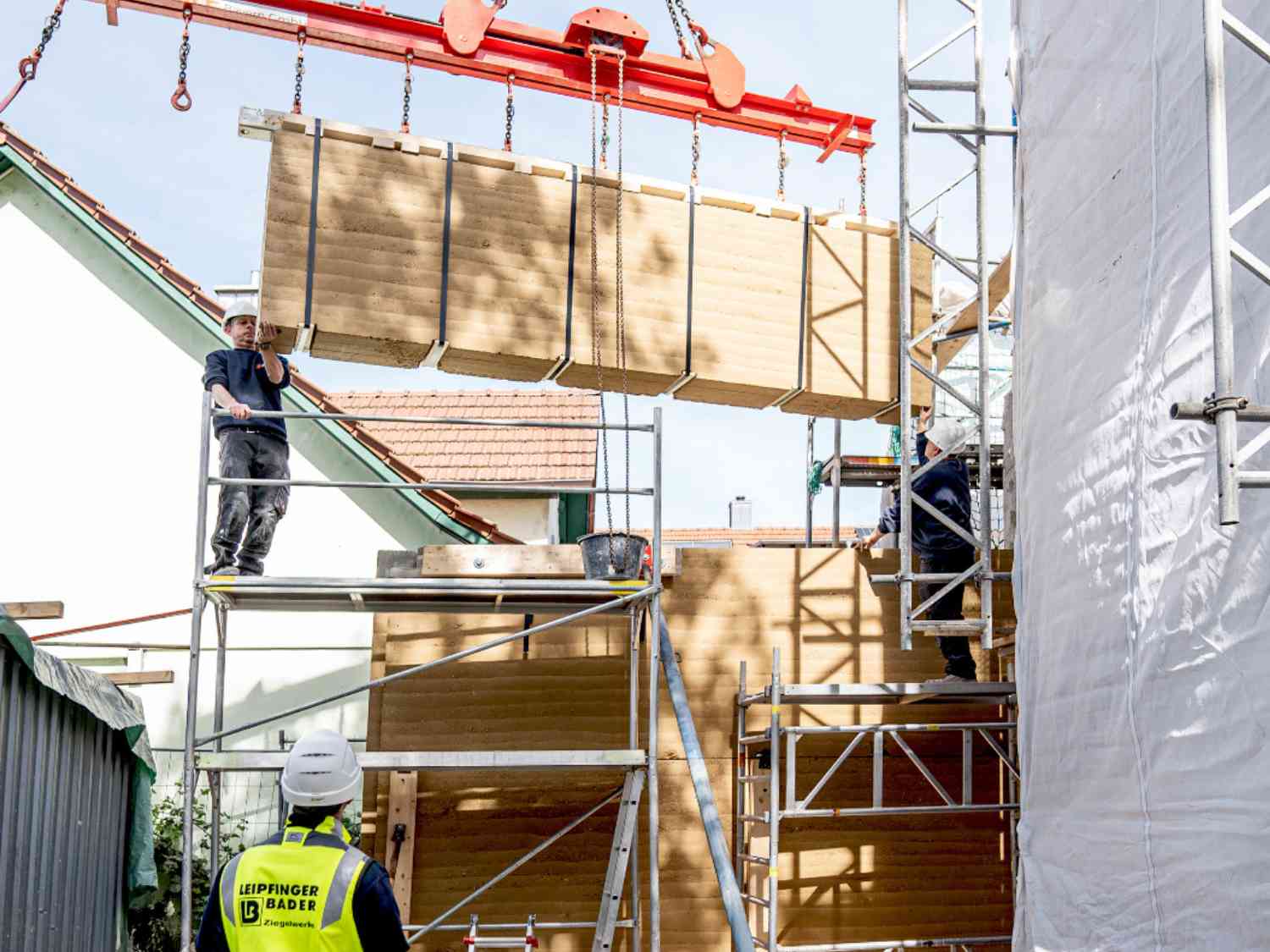 Zwei Bauarbeiter montieren eine hängende Stampflehmwand von Leipfinger-Bader an einem Gerüst auf einer Baustelle.