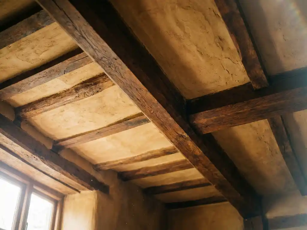 Dicke Holzbalkendecke mit natürlichem Lehm zwischen dunklen Balken, warmes Honigholz und erdige Lehmoberflächen im Tageslicht